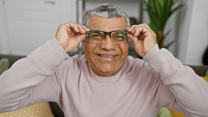 A smiling mature man adjusts his glasses in a cozy living room, exuding warmth and contentment.