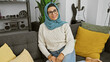 © Krakenimages.com - A smiling mature woman wearing a hijab sitting comfortably in a modern living room on a gray couch with colorful pillows.
