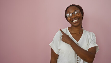 Sticker - African american woman smiling and pointing while standing against an isolated pink background, wearing glasses and a white top, showcasing a cheerful expression and positive attitude.