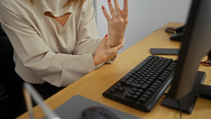 A woman in an office holds her wrist in discomfort, indicating potential wrist pain or carpal tunnel syndrome, while sitting at a computer desk indoors.