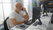 © Krakenimages.com - Man working on computer in office, bald with grey hair and beard, seated indoors while talking on phone, smiling in modern workplace with windows, plants, and office supplies on desk