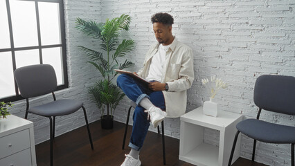 Young african american man with a beard reading a clipboard while seated in an indoor waiting room with a modern interior and minimalistic decor.