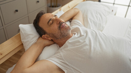 Handsome middle-aged hispanic man relaxing on a white bed in a cozy bedroom with his hands behind his head, enjoying a moment of peace.