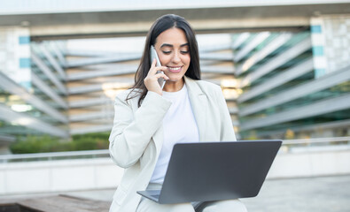 Poster - A woman engaged in a phone call while using her laptop, smiling in an outdoor workspace surrounded by modern architecture.