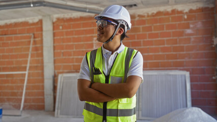 Wall Mural - Young chinese man with crossed arms wearing a hard hat and safety vest standing indoors on a construction site with red brick walls and equipment in the background