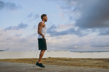 Wall Mural - Side view young strong sporty athletic fit sportsman man wears sports clothes jumping on skipping rope warm up train at sunrise sun dawn over sea beach outdoor on pier seaside in summer day morning