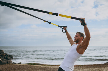 Wall Mural - Profile young strong sporty athletic toned fit sportsman man wear sports clothes use trx loops do exercise warm up training at sunrise sun dawn over sea beach outdoor on seaside in summer day morning.