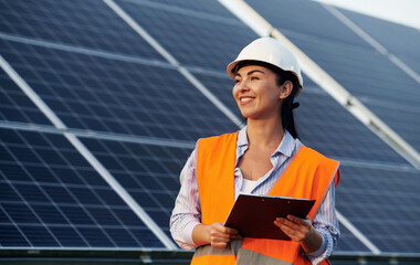 Notepad in hands. Female worker engineer in uniform is near solar panels