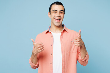 Wall Mural - Young smiling happy cheerful middle eastern man he wear pink shirt white t-shirt showing thumb up like gesture isolated on plain pastel light blue cyan background studio portrait. Lifestyle concept.