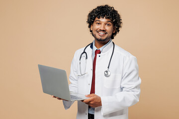 Wall Mural - Young happy Indian doctor man wear white medical gown suit work in hospital clinic office hold use laptop pc computer isolated on plain beige background studio portrait. Healthcare medicine concept.