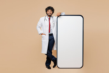 Wall Mural - Full body young Indian doctor man wear white medical gown suit work in hospital clinic office stand near big huge blank screen mobile cell phone isolated on plain beige background. Healthcare concept.