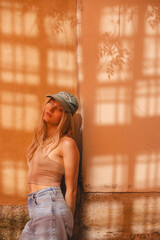 Fashion portrait pretty sensual woman posing in the city near wall, street fashion. Girl wear beige top, green cap and blue jeans. Sunny light. Dreaming thoughtful girl in casual outfit look calm.