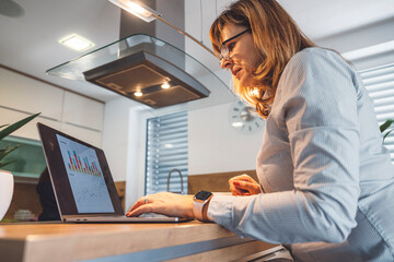 A woman sitting at a kitchen island, looking stressed while using a laptop. She has long brown hair and wears a light blue shirt. A small plant and a cup are on the counter.