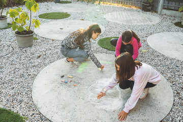 Wall Mural - Three children drawing with chalk on a concrete surface in a garden. The setting features gravel and circular stone patterns, with green plants in pots around them. The sunlight casts a warm glow on t