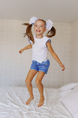 Cheerful little girl with ponytails and white bows jumping on the bed.