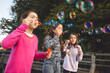 © VisualProduction - Three girls blowing bubbles outdoors, surrounded by trees. They are wearing colorful sweaters and smiling, enjoying a playful moment. The bubbles are floating in the air, reflecting sunlight.