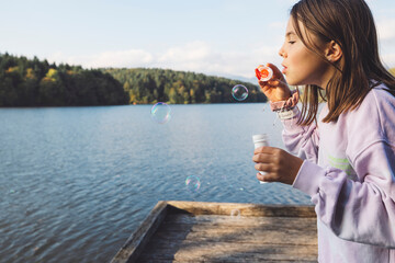 Wall Mural - A young girl blowing bubbles by a serene lake, surrounded by trees. She wears a light purple sweater and has a joyful expression. The bubbles float in the air, reflecting sunlight.