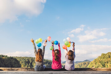 Wall Mural - Three girls sitting on a wooden dock, facing away, holding colorful balloons. They are enjoying a sunny day with a scenic view of trees and mountains in the background.