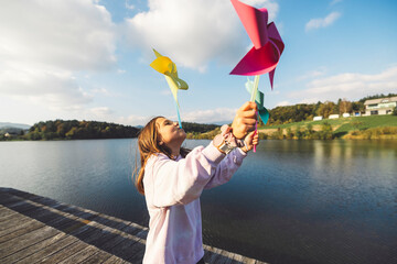 Wall Mural - A young girl joyfully playing with colorful pinwheels by a lake. She stands on a wooden pier, holding the pinwheels up to her face, with a serene landscape in the background and a clear blue sky.