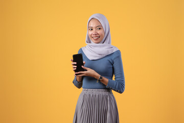 Wall Mural - happy asian indonesian muslim woman holding smart phone and showing blank screen on isolated yellow background