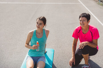 A young woman sitting on an exercise mat, holding a water bottle, while an older woman embraces her from behind, both smiling. They are outdoors on a sunny day, suggesting a fitness or training sessio