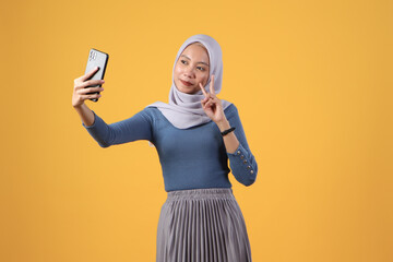 Wall Mural - happy asian indonesian muslim woman holding smart phone giving victory finger gesture on isolated yellow background