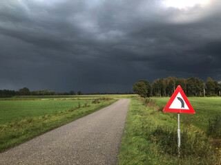 Countryroad near Uffelte with dark rainclouds in the back. Meadows at the Turfvenenweg. Drenthe Netherlands. Traficsign. Curve coming up. Warning.