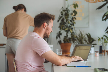 A man working on a laptop in a bright, modern workspace filled with plants. He is focused on the screen, typing on the keyboard, with a notebook and a glass of water nearby.