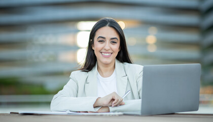 Wall Mural - A woman sits at a table, smiling as she works on her laptop in an outdoor setting during the afternoon.