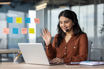 Wall Mural - Latin American woman with headset phone using laptop for video call, female online customer support worker waving hand greeting customer, online meeting with colleagues remotely.