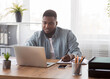 © Prostock-studio - Young black businessman using laptop and taking notes at worklace in modern office,free space