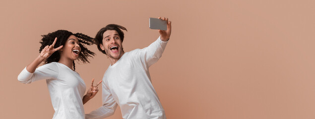 Wall Mural - Selfie Fun. Millennial Interracial Couple Jumping And Taking Self-Portrait, Posing Together Over Yellow Background, Afro Girl Showing Peace Gestures