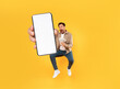 © Prostock-studio - A man leaps in the air while holding a handful of money in one hand and a smartphone displaying a blank screen in the other, set against a vibrant yellow backdrop, showcasing enthusiasm and surprise.