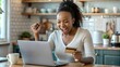 © Htet Wai Phyo - Happy excited young African woman customer winner holding credit card using laptop computer in kitchen celebrating money win receiving cashback getting online bonus for shopping sitting at home