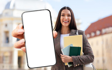 A young woman smiles broadly while holding a blank smartphone in one hand and several colorful files in the other, standing in front of an attractive modern building on a sunny day.