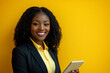 © Volodymyr - Young Black businesswoman holding a touchpad in an office, smiling brightly at the camera, against a yellow background.