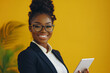 © Volodymyr - Young Black businesswoman holding a touchpad in an office, smiling brightly at the camera, against a yellow background.