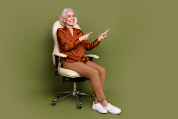 Full size photo of pretty retired lady sit office armchair point look empty space wear brown formalwear isolated on khaki color background