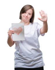 Young adult woman with down syndrome holding red heart card over isolated background with open hand doing stop sign with serious and confident expression, defense gesture