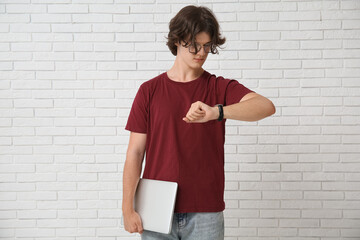 Male programmer in eyeglasses with laptop looking at wristwatch on white brick background