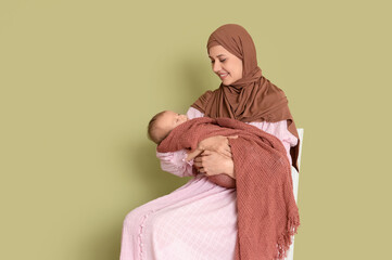 Wall Mural - Happy Muslim mother in hijab with cute little baby sitting on chair against green background