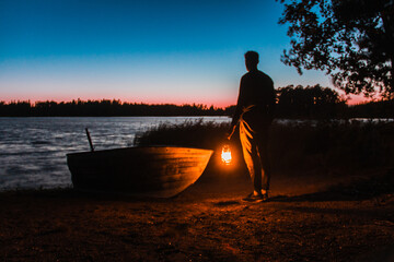 A man holding a storm lamp by an old row boat at sunset. Finnish archipelago.