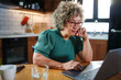 © lordn - A mature woman with curly gray hair and glasses works on a laptop in a cozy home kitchen. The setting is relaxed, with natural light coming in through the window, creating a productive atmosphere