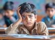 © Boy photographer - boy sitting at a desk in a classroom, holding his head with his hand and looking distressed while doing homework. The teacher and other students are blurred in the background.