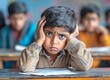© Boy photographer - boy sitting at a desk in a classroom, holding his head with his hand and looking distressed while doing homework. The teacher and other students are blurred in the background.