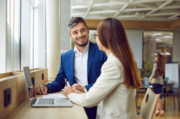 Wall Mural - Two happy smiling company employees man and woman sitting with laptop having discussion, talking about new job projects or startups. Business people working together in modern office.