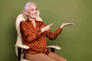 Poster - Photo of attractive senior woman sit armchair look point hold empty space dressed brown formalwear isolated on khaki color background