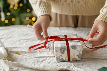 Wall Mural - Wrapping stylish christmas gift, winter holidays preparation. Hands in cozy sweater holding gift box and festive paper on rustic table on background of decorated christmas tree with lights