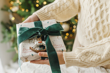 Wall Mural - Merry Christmas and Happy Holidays. Hands in sweater holding modern christmas gift box with green ribbon and vintage toy on background of stylish christmas tree with lights.