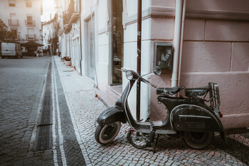 Vintage black scooter parked against an old city wall on a cobblestone street, with soft morning light creating a nostalgic urban atmosphere in a European setting; Leiria, Portugal